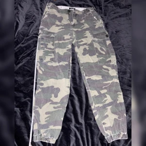 Baggy cargobyxor i camouflage med resår fashionnova S-XS - Säljer ett par cargobyxor i klassiskt khaki grönt camouflage-mönster med flera fickor och resår vid bensluten. Byxorna har dragkedja och knapp framtill samt hällor för bälte. Perfekta för en avslappnad streetstil! 