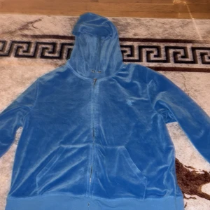 Blå velour hoodie från Juicy Couture - Blå hoodie i mjuk velour från Juicy Couture med broderad JC-logga på bröstet. Tröjan har huva med snörning, dragkedja framtill och två fickor. Perfekt för chill dagar och har en riktigt skön känsla mot huden.