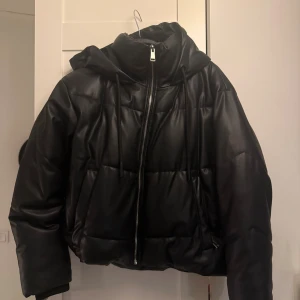 Svart pufferjacka från Zara - Svart pufferjacka från Zara med snygg quiltad design och huva. Jackan har dragkedja framtill och snörning vid halsen. Materialet är syntet med skinnlook som ger en cool vibe. Perfekt till den kalla vintern som kommer. slutsåld på zara