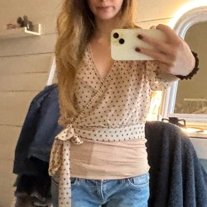 Beige omlottblus med prickigt mönster - Säljer en beige omlottblus med svart prickigt mönster och knyt i midjan. Blusen har v-ringad hals och korta ärmar, vilket ger en chill och feminin vibe. Perfekt att styla med jeans eller kjol för en snygg look.