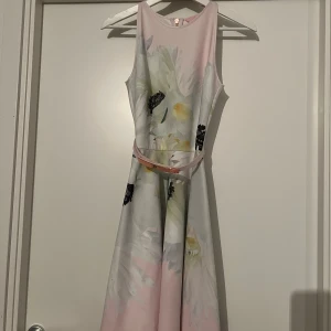 Blommig ljus klänning från Ted Baker - Superfin klänning från Ted Baker med ljusrosa och vita toner, stora blommor och ett smalt bälte med roségulddetalj. Klänningen är ärmlös, har rund hals och dragkedja bak. Materialet är mjukt och har en lätt glansig finish, perfekt för vår och sommar. USA storlek 0 motsvar xs. 