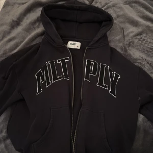 Svart hoodie från Multiply Apparel - Svart hoodie från Multiply Apparel med vit broderad text på bröstet. Tröjan har dragkedja framtill, huva och två fickor. Perfekt för en chill och avslappnad stil. Klassisk passform och mjukt material.