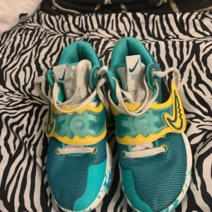 Turkosa Nike sneakers med gula detaljer - Säljer ett par unika Nike basketskor i turkos med coola gula detaljer och vit sula med blått mönster. Skorna har snörning och en vågig design på sidan med ett stort gult Swoosh. Ovandelen är i syntetmaterial och mesh för bra ventilation. Perfekt för dig som gillar färgstarka sneakers.