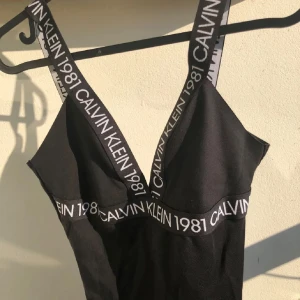 Svart boddy Calvin Klein 1981 - Svart boddy från Calvin Klein med breda axelband och resår under bysten, går att använda som baddräkt oxå men de är tyg-material. Den har vit logotyptext 'CALVIN KLEIN 1981'. v-ringning både fram och bak. Perfekt för stranden eller poolen. Mycket stretch i