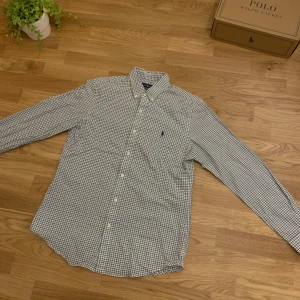 Rutig slim fit skjorta från Ralph Lauren - Riktigt snygg rutig skjorta från Ralph Lauren i slim fit-modell. Skjortan är vit med blå och gröna rutor, har klassisk krage, långa ärmar och knappar framtill. Ikoniska Polo-loggan broderad på bröstet. Perfekt för dig som gillar stilrena och fräscha plagg.