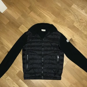 Svart Moncler pufferjacka med stickade ärmar - Svart pufferjacka från Moncler med stickade ärmar och klassisk logga på vänster arm. Jackan har quiltad front, två dragkedjeförsedda fickor och hel dragkedja framtill. Materialet är en mix av polyester och bomull, vilket gör den både mjuk och varm.