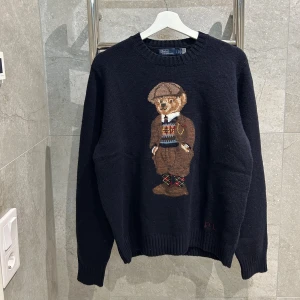 Svart stickad tröja Polo Bear Ralph Lauren - Marinblå stickad tröja från Polo Ralph Lauren med klassisk Polo Bear-motiv framtill. Tröjan har rund hals, långa ärmar och ribbade muddar. RL-broderi vid nederkanten ger extra detalj. Använd cirka 2 gånger. Skick 10/10