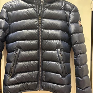 Mörkblå Colmar pufferjacka med huva - Snygg mörkblå pufferjacka från Colmar med huva och två fickor med dragkedja. Jackan har quiltad design och en klassisk logga på ärmen. Perfekt för kalla dagar. Endast använd en vinter, är i mycket fint skick. Liten i storlek.