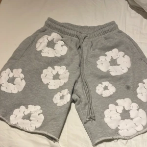 Grå shorts med vita tryck från Denim Tears - Säljer ett par grå shorts från Denim Tears med unika vita tryck i form av blommor över hela plagget. De har elastisk midja med snörning och råa kanter vid bensluten. Perfekta för chill dagar och har en riktigt skön street vibe.