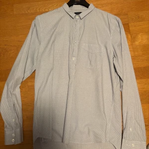 Rutig henley-skjorta från Velour M - Snygg henley-skjorta från Velour i vitt med tunna blå rutor. Skjortan har lång ärm, bröstficka och knappar framtill. Tillverkad i mjuk bomull och passar perfekt för en clean och avslappnad look.