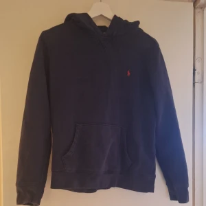 Mörkblå hoodie från Polo Ralph Lauren - Säljer en mörkblå hoodie från Polo Ralph Lauren med klassisk huva och känguruficka. Tröjan har en liten röd broderad logga på bröstet och är tillverkad i mjuk bomullsmix. Perfekt för chill dagar eller när du vill ha en clean och stilren look.