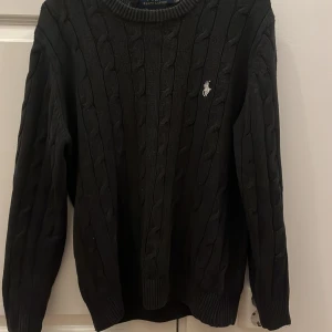 Svart stickad tröja Polo Ralph Lauren - Svart stickad tröja från Polo Ralph Lauren med klassisk rund halsringning och broderad logga på bröstet. Tröjan har långa ärmar och ribbade muddar för en snygg passform. Perfekt för dig som gillar stilrena och tidlösa plagg.