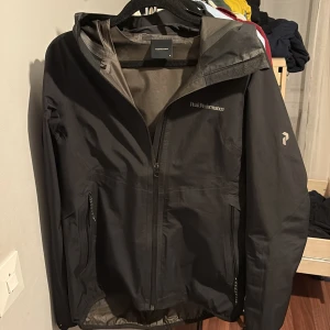 Svart vindjacka Peak Performance M - Svart vindjacka från Peak Performance i storlek M. Jackan har huva, hel dragkedja framtill och två sidofickor med dragkedja. Diskret logga på bröstet och ärmen. Perfekt för blåsiga dagar och outdoor-aktiviteter.