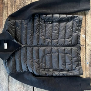 Svart pufferjacka från Moncler - Svart pufferjacka från Moncler med stickade ärmar och quiltad front. Jackan har två sidofickor med dragkedja och en bröstficka med dragkedja. Moncler-logga på vänster ärm. Snygg, stilren och perfekt för kyliga dagar. Köpt för ett par sen från Moncler och har använt den ungefär 10-15 gånger riktigt bra skick på den. Kvitto kommer privat 