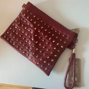 Röd kuvertväska med nitar och tofs - Snygg röd kuvertväska med guldfärgade nitar och matchande tofs. Väskan har avtagbar axelrem och handledsrem i flätad stil. Tillverkad i syntetmaterial med dragkedja upptill. Perfekt accessoar för att lyfta din outfit.