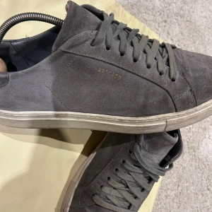 Grå mocka sneakers Axel Arigato - Snygga grå sneakers från Axel Arigato i mjuk mocka med diskret guldfärgad logga på sidan. Skorna har rund tå, platt vit sula och grå snörning. Baksidan har svart skinnpanel och Arigato-tryck på hälen. Ingår en påse! Är öppen för bud!