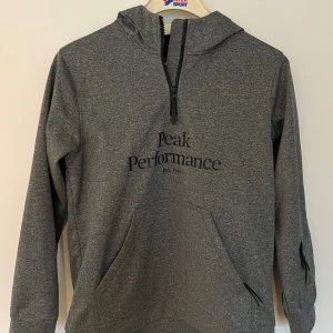 Grå hoodie från Peak Performance - Snygg grå hoodie från Peak Performance med svart logga på bröstet. Tröjan har huva, halv dragkedja och en stor magficka. Perfekt för chill dagar eller när du vill ha en sportig look. Långärmad och i mjukt material.