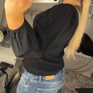 Svart blus med knappar och puffärm - Säljer en svart blus med rundad halsringning och knappar framtill. Blusen har långa puffärmar med resår vid ärmslut och är lätt croppad i modellen. Perfekt att styla med jeans för en cool och enkel look.
