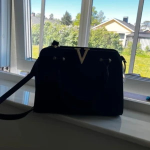 Svart handväska med guldiga detaljer - Snygg svart handväska med strukturmönster och guldigt V framtill. Väskan har både handtag och justerbar axelrem, samt dragkedja och extra fack på baksidan. Perfekt för dig som vill ha en stilren och trendig accessoar.