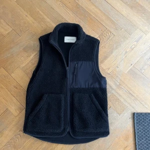 Svart fleeceväst från A Day's March - Svart fleeceväst med hög krage och två stora fickor framtill. Västen har detaljer i slät polyester på bröstet med en praktisk dragkedja. Perfekt för lager-på-lager och chill stil. Stängs med dragkedja framtill. Kontakta för fler frågor 