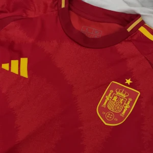 Spaniens landslagströja Adidas röd L - Säljer en röd fotbollströja från Spaniens landslag. Tröjan har gula detaljer, landslagets emblem på bröstet och klassiska Adidas-ränder på axlarna. Materialet är lätt och andas, perfekt för match eller träning.