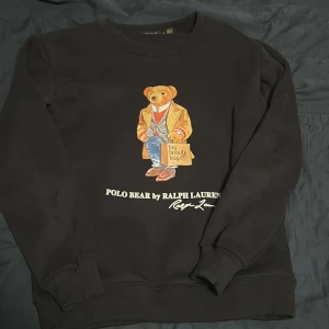 Svart sweatshirt Polo Bear Ralph Lauren - Svart sweatshirt från Polo Ralph Lauren med ikoniska Polo Bear-trycket framtill. Tröjan har rund hals, långa ärmar och ribbade muddar. Perfekt för dig som gillar streetstyle och vill ha något klassiskt men ändå lekfullt.