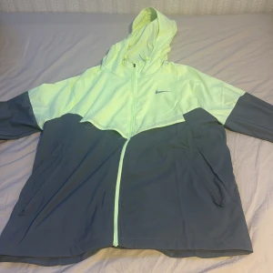 Nike vindjacka i ljusgrön och grå - Nike vindjacka med huva i ljusgrön och grå färgblock. Jackan har hel dragkedja, snörning vid huvan och två sidofickor med dragkedja. Materialet är lätt och syntetiskt, perfekt för löpning och utomhusaktiviteter. Snygg Nike-logga på bröstet och meshfoder på insidan. 