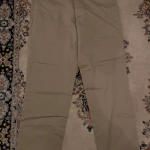 Beiga chinos från Dockers, storlek 36x34 - Snygga beiga chinos från Dockers i klassisk rak modell. Byxorna har två bakfickor med knapp och är gjorda i mjukt bomullsmaterial. Perfekt passform och stilren look som funkar till många olika outfits. Taggarna sitter kvar.