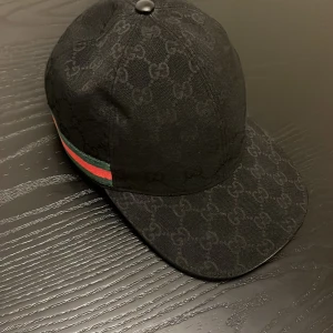 Svart Gucci keps med GG-mönster - Snygg svart keps från Gucci med klassiskt GG-monogram över hela kepsen och en grön-röd rand på sidan. Justerbar rem baktill i skinn och insida med Gucci-tag. Perfekt för dig som vill ha en lyxig streetstyle-accessoar.