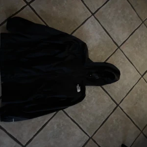 Svart vindjacka från The North Face - Svart vindjacka från The North Face med huva och dragkedja framtill. Jackan har en stilren look med logotypen tryckt på bröstet. Perfekt för blåsiga dagar och enkel att matcha med olika outfits. Långärmad och tillverkad i lätt syntetmaterial.