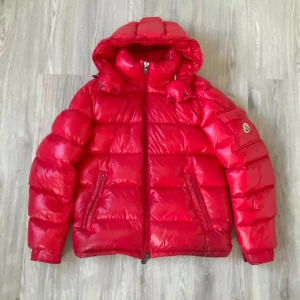 Röd Moncler Maya - Säljer en fet röd Moncler Maya pufferjacka i storlek M. Jackan har en glansig yta, huva, bröstficka med Moncler-logga och bruna detaljer på insidan. Fylld med dun och har dragkedja framtill samt två sidofickor med zip. Perfekt för kalla dagar. Årsmodell 2022