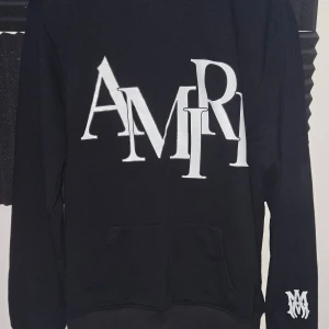 AMIRI hoodie - Svart hoodie från AMIRI med stor vit logotyp framtill och en mindre logga på ärmen. Tröjan har huva, känguruficka och långa ärmar. Materialet är mjuk bomull som känns skönt mot huden. Perfekt för dig som gillar streetwear och vill sticka ut med en fet design.