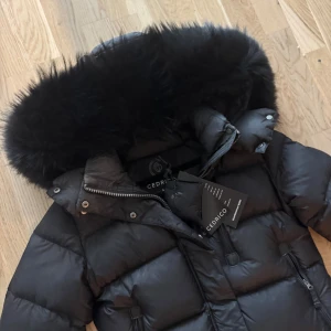 Svart dunjacka med pälskrage från Cedrico  - Snygg svart dunjacka från Cedrico med en fluffig pälskrage. Jackan har en lång design med dragkedja och knappar framtill. Perfekt för kalla vinterdagar med sin varma och skyddande stil. Modellen är Stella Long. Jackan är knappt använd en hel vinter, jätte fint skick. Kan dock behöva tvättas. Köpt på Johnells för 7499kr. Alla lappar kvar 