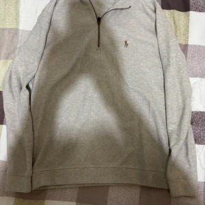 Beige half zip tröja från Polo Ralph Lauren - Snygg beige långärmad tröja från Polo Ralph Lauren med half zip och klassisk broderad logga på bröstet. Tillverkad i 100% bomull, perfekt för dig som gillar stilrena och bekväma plagg. Passar dig som vill ha en enkel men ändå ikonisk look.