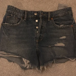 Högmidjade jeansshorts med fransar - Säljer ett par mörkblå högmidjade jeansshorts med slitna detaljer och fransig kant. Klassisk femficksmodell med knäppning framtill och råa benslut för en cool, avslappnad vibe. Perfekta för sommaren och festivaler.