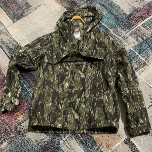 Camouflage vindjacka från Carhartt - Säljer en grön och brun camouflage vindjacka från Carhartt i storlek XS. Jackan har huva, dragkedja framtill och är tillverkad i 100% nylon. Perfekt för dig som gillar streetwear och vill sticka ut med ett fett mönster.