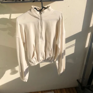 Beige half zip hoodie  - Snygg beige/vit croppad hoodie med half zip framtill och broderad logga på bröstet. Tröjan har lång ärm, känguruficka och elastisk midja jätte skön lite tunnare material o luftig