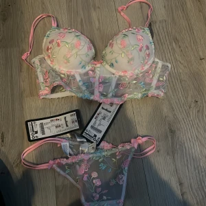 Blommig korsett från New Yorker - Superfin korsett från New Yorker i transparent mesh med broderade blommor i rosa, blått och grönt. Den har ljusrosa axelband och små rosetter framtill. Perfekt för dig som gillar romantiska detaljer och färgglada inslag. Säljer med matchande trosa! Storlek XS i båda 75A