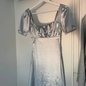 Silvrig kort klänning från H&M  - Supersnygg silvrig klänning från H&M  i kort modell med puffiga korta ärmar och fyrkantig urringning. Materialet är glansigt och känns lyxigt, perfekt för dig som gillar att sticka ut. Passformen är normal och klänningen har en enkel, stilren design.