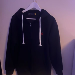 Svart hoodie från Polo Ralph Lauren - Svart hoodie från Polo Ralph Lauren med vit snörning och dragkedja framtill. Klassisk röd logga broderad på bröstet. Tröjan har huva och långa ärmar, perfekt för chill dagar. Mjuk bomullskänsla och normal passform.