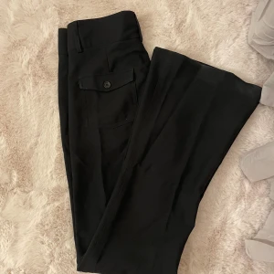Lågmidjade bootcut kostymbyxor  - Svarta kostymbyxor med bootcut och en snygg ficka med knapp bak. Byxorna har klassisk look och är perfekta för dig som gillar en stilren och trendig vibe. Defekt på bild två, syns knappt. Sitter som S.