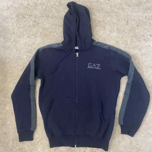 Mörkblå EA7 hoodie med dragkedja - Snygg mörkblå hoodie från EA7 Emporio Armani med gråa detaljer längs ärmarna. Tröjan har huva med snörning, hel dragkedja framtill och två stora fickor. Perfekt för chill eller träning, stilren logga på bröstet och mjukt bomullsmaterial.