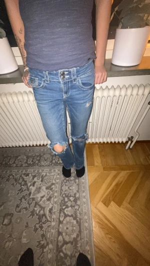 Low waist bootcut jeans med hål - Säljer ett par blå jeans med utsvängda ben och slitna detaljer vid knäna. Jeansen har dubbla knappar i midjan, tydliga kontrastsömmar och klassiska fickor fram och bak. Perfekta för dig som gillar en avslappnad och trendig look.