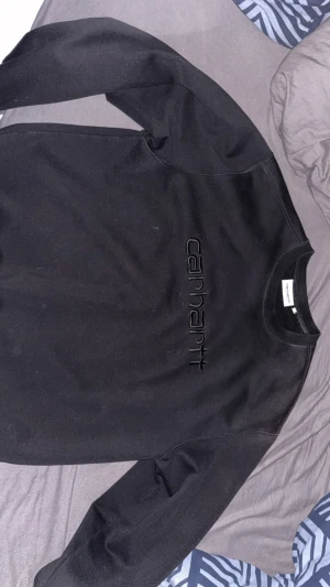  sweatshirt  Carhartt - Svart sweatshirt från Carhartt med broderad logga framtill. Tröjan har rund hals, ribbade muddar och är tillverkad i mjuk bomull med insida i fleece. Perfekt för dig som gillar streetwear och vill ha en clean look.