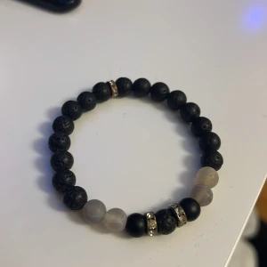 Svart och grått pärlarmband - Stilrent armband med svarta lavapärlor och matta grå pärlor, dekorerat med två guldfärgade detaljer med små stenar. Perfekt accessoar för att lyfta din outfit med en cool och modern vibe.