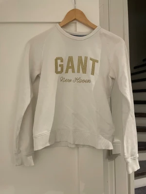 Vit sweatshirt från GANT med guldtext - Vit sweatshirt från GANT med snyggt broderad guldtext 'GANT New Haven' på bröstet. Tröjan har rund hals, ribbade muddar och är långärmad. Perfekt för dig som gillar klassisk och stilren design med en touch av lyx.