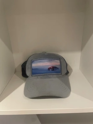 Grå truckerkeps från Revenite - Snygg grå truckerkeps från Revenite med mesh på baksidan och justerbar snapback. Framtill finns en cool patch med bilmotiv i blå och rosa toner. Kepsen har mjuk, mockaliknande front och logotyp på insidan. Perfekt för dig som gillar streetstyle.
