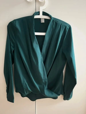 Grön omlottblus från H&M - Snygg mörkgrön omlottblus från H&M med långärmad design och djup v-ringning. Blusen har ett elegant omlottfall framtill och är tillverkad i mjuk och slät polyester som faller fint. Perfekt att styla med jeans eller kjol för en trendig look. Tyvärr har blusen ett par defekter vid sidan