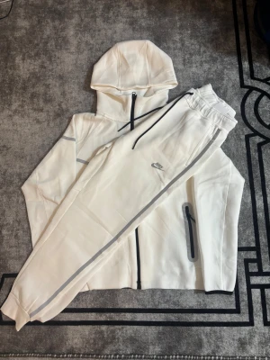 Vit Nike reclective tech tracksuit - Säljer ett par vita mjukisbyxor från Nike med snygga reflexdetaljer längs benen och dragkedjeförsedda fickor. Byxorna har elastisk midja med snörning och är tillverkade i mjukt material som passar perfekt för chill eller träning. Nike-logga på låret för extra stilpoäng.
