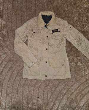 Beige quiltad jacka från Barbour - Snygg beige quiltad jacka från Barbour International med klassisk krage, guldfärgade knappar och dragkedja. Jackan har flera fickor med lock och Barbour-logga på bröstet. Perfekt för dig som gillar stilren och tidlös design. Lite trådar på sista bild vid ficka annars väldigt fin 
